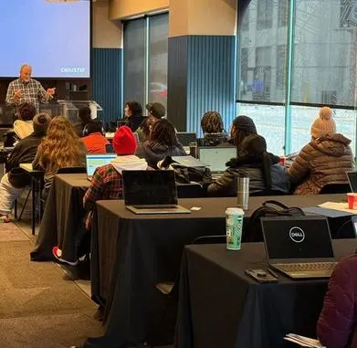 People seated at tables with their laptops listening to an instructor at the front of the room.