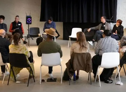 A group of people seated in a circle of chairs talking.