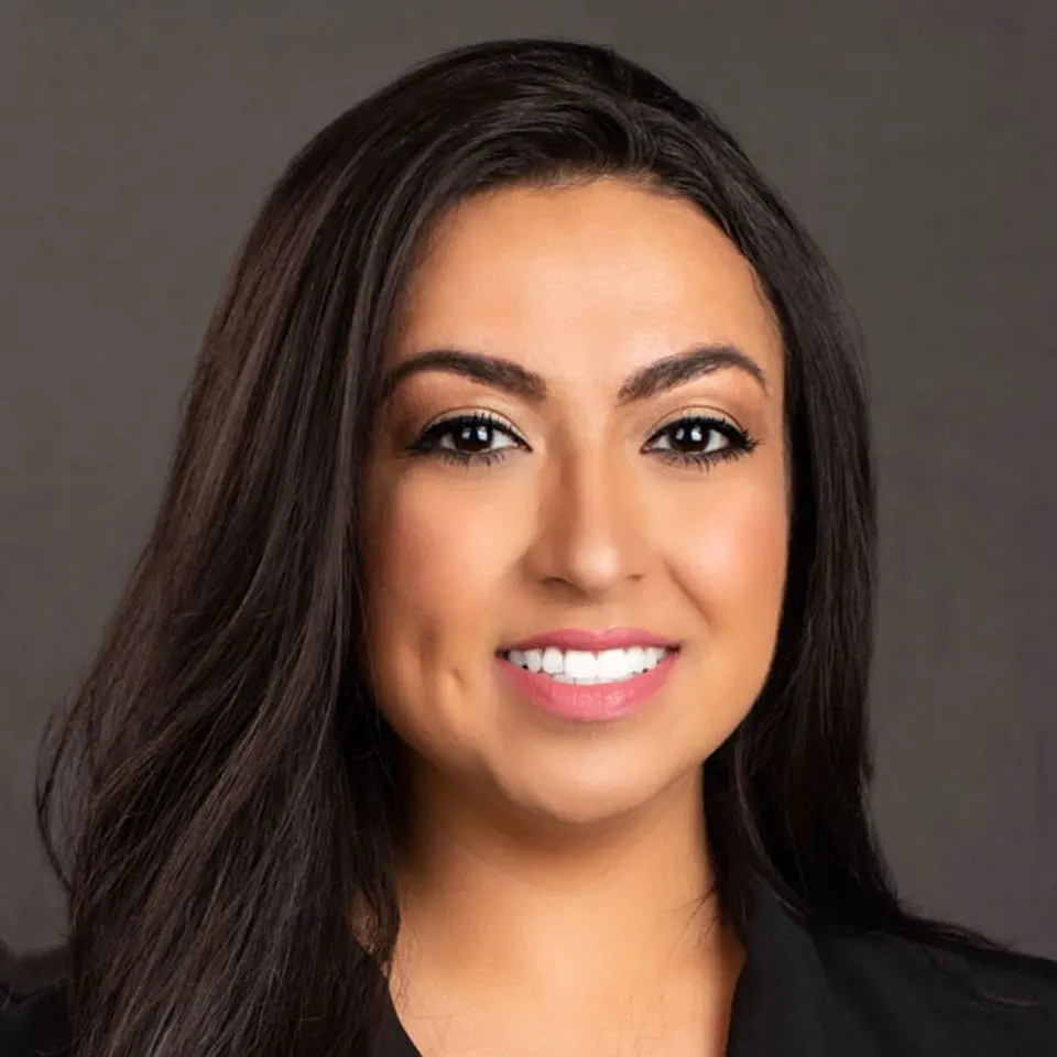 Woman with long dark hair and a black blazer smiling in front of a plain dark background.