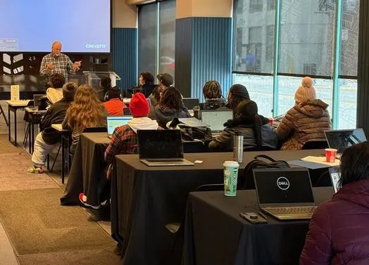 People seated at tables with their laptops listening to an instructor at the front of the room.