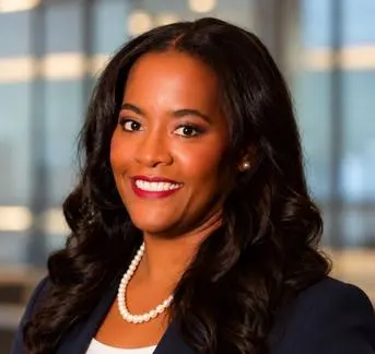 A black woman with long wavy dark hair wearing a navy blazer, white shirt and a pear necklace.