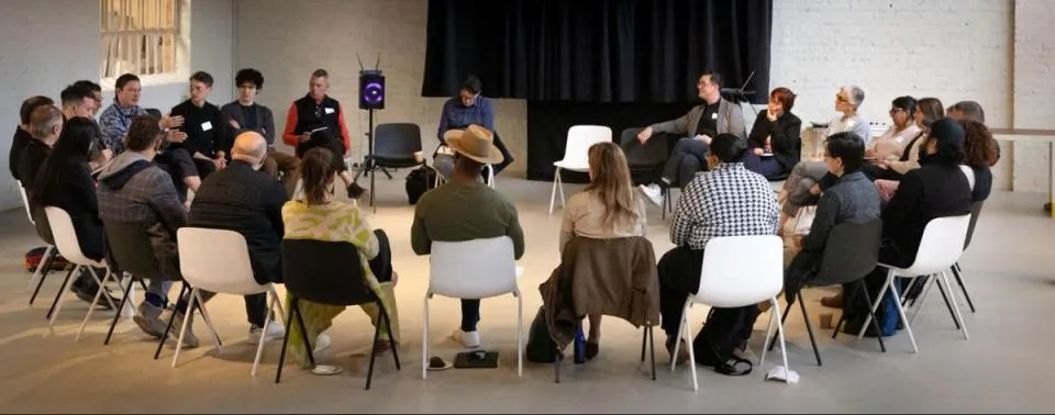 A group of people seated in a circle of chairs talking.